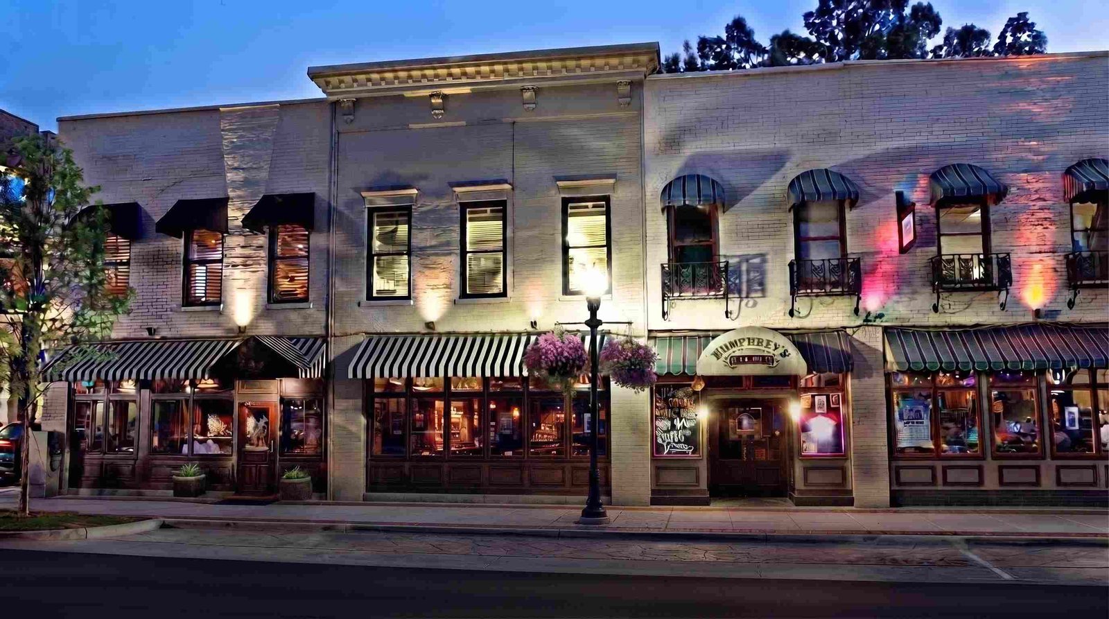 Historic brick storefronts lit at dusk in Huntsville's Twickenham Historic District, with period streetlamp and hanging flower baskets